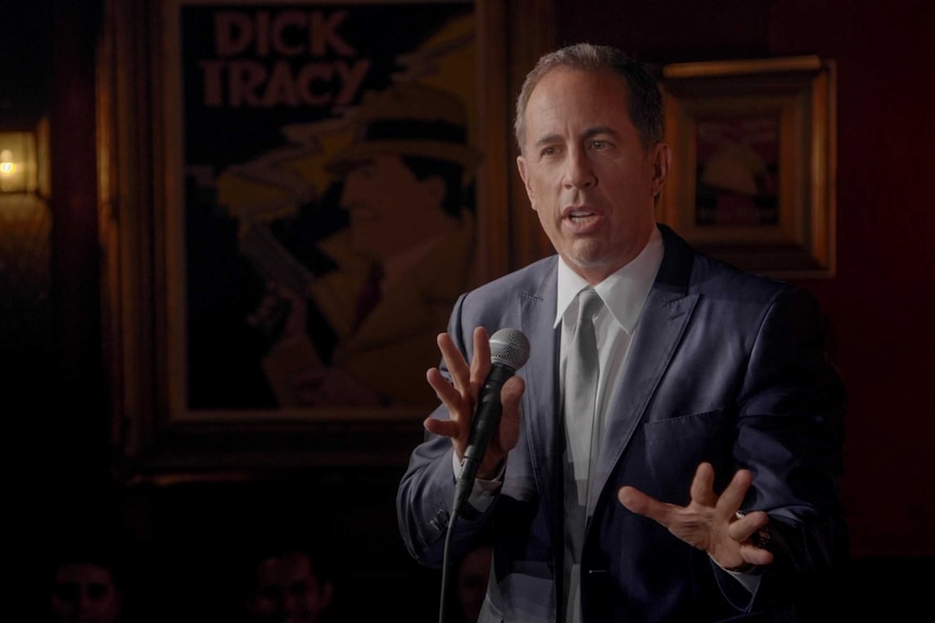 Jerry Seinfeld performs on stage at a comedy club.