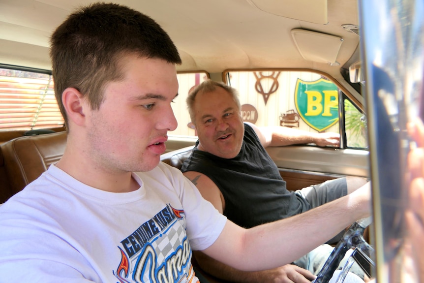 Young man in white shirt nearest to camera looks down as man in black top in seat next to him looks across.