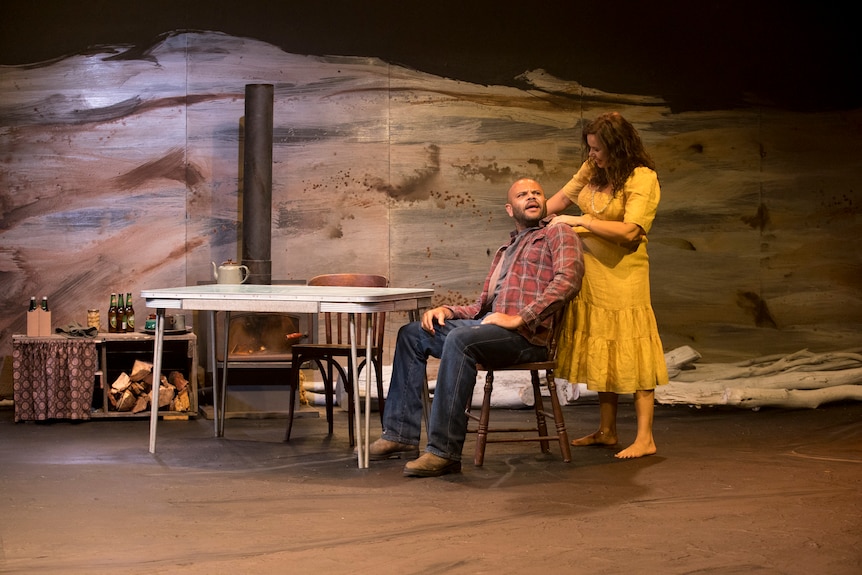 A male and female actor on a stage. The man sits at a table while the woman stands behind him, her hands on his shoulders.