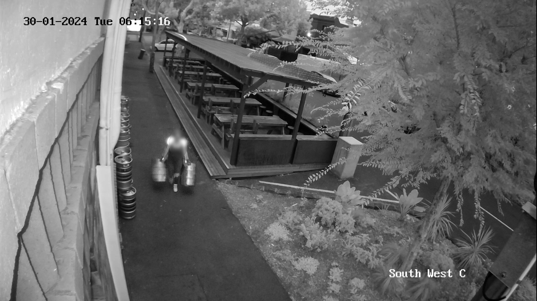 Black and white CCTV footage of a woman wearing high-vis holding two kegs walking past outside of pub