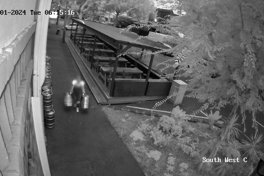 Black and white CCTV footage of a woman wearing high-vis holding two kegs walking past outside of pub
