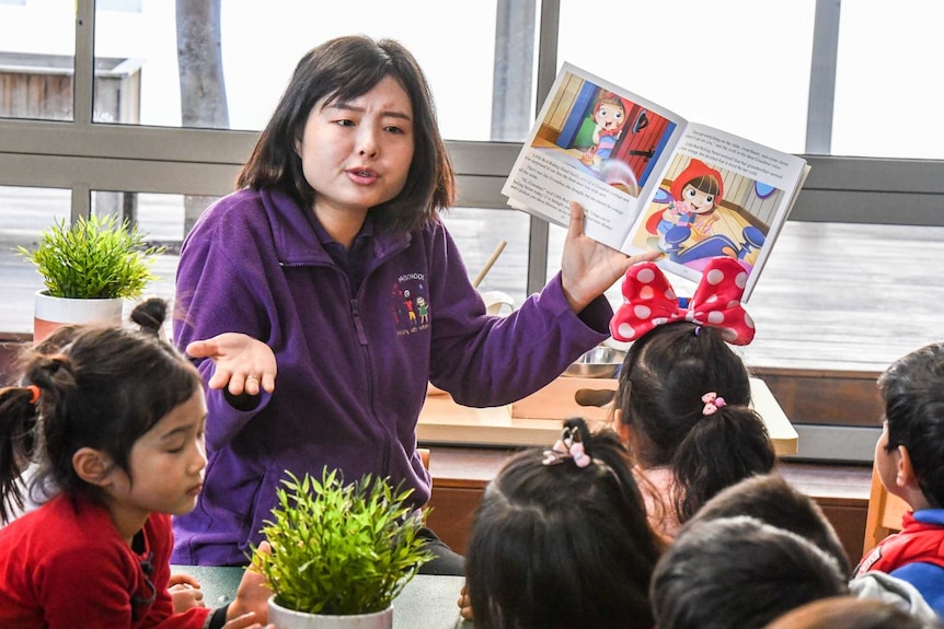 a woman in a purple jumper holds up a picture book with children infront of her