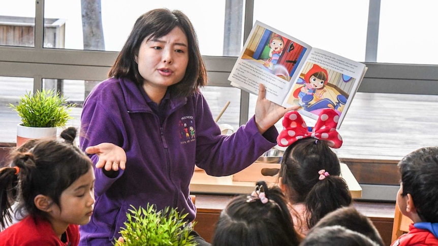 a woman in a purple jumper holds up a picture book with children infront of her