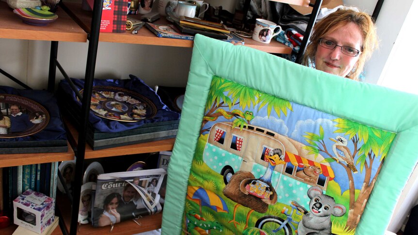 Woman stands holding quilt containing cartoon images of emu, koala and kangaroo.