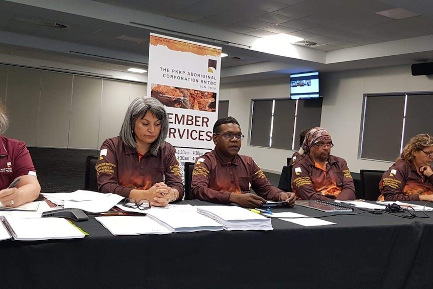 Five people sitting at a long table, wearing a brown uniform, with lots of notes and papers on the table, in a conference room