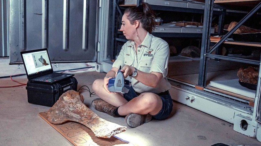 Samantha Beeston sits on the floor of a shed holding a scanning device over a large dinosaur bone. 