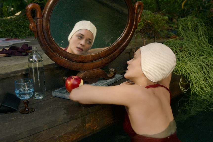 Marion Cotillard in maroon bathers rests her head on the steps of a riverbank and looks longingly at her reflection in a mirror.