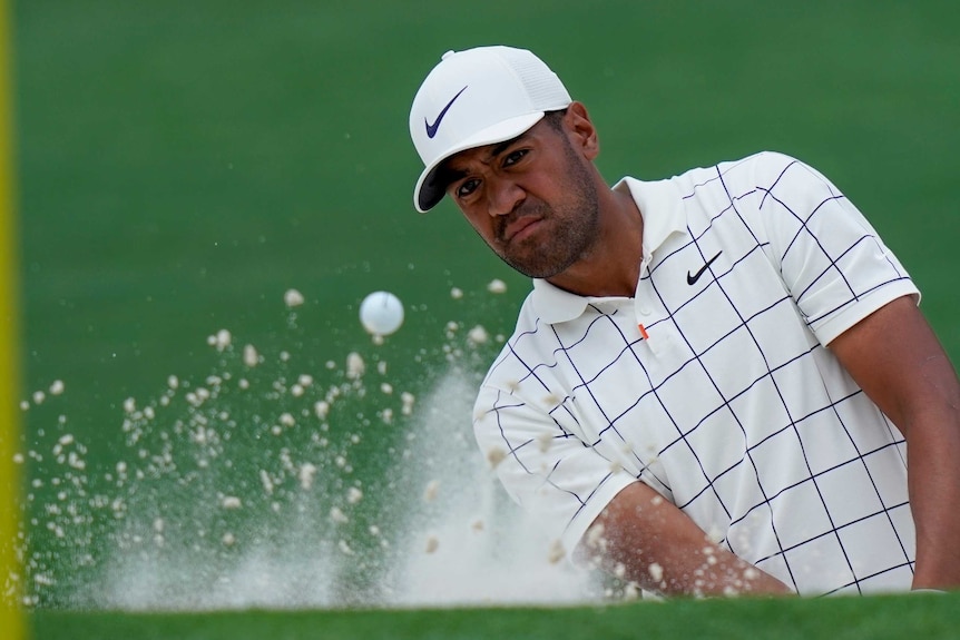 Tony Finau watches the ball head towards a yellow pole in the foreground as sand sprays up around him