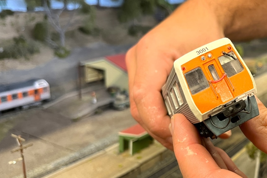 A pair of hands hold a model of a silver and orange Adelaide suburban train.