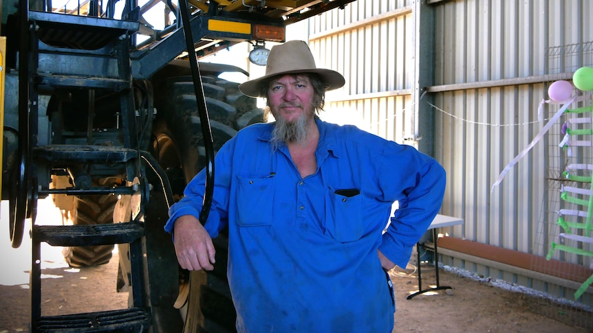 A man with a beard in a blue shirt learning on a tractor. 