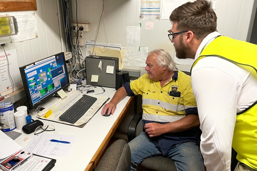 An older man with grey hair and a young man with glasses, brown beard, both wear high-vis, look at an computer in a room.