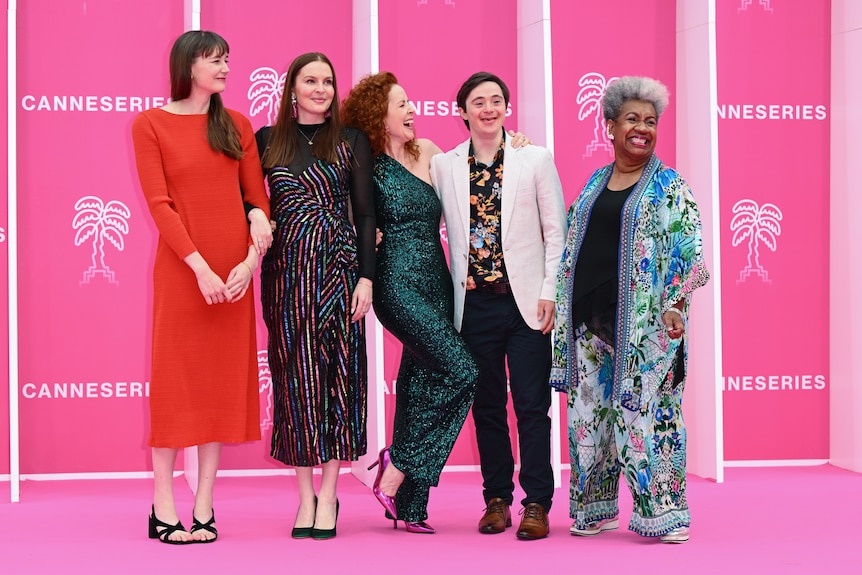 Three white women, one white man and one Pasifika woman are dressed in glamourous formal-wear on a film festival pink carpet.
