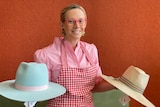 Woman in pink shirt and red apron smiles holding a blue hat and straw hat 