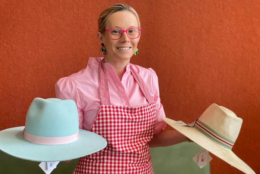 Woman in pink shirt and red apron smiles holding a blue hat and straw hat 