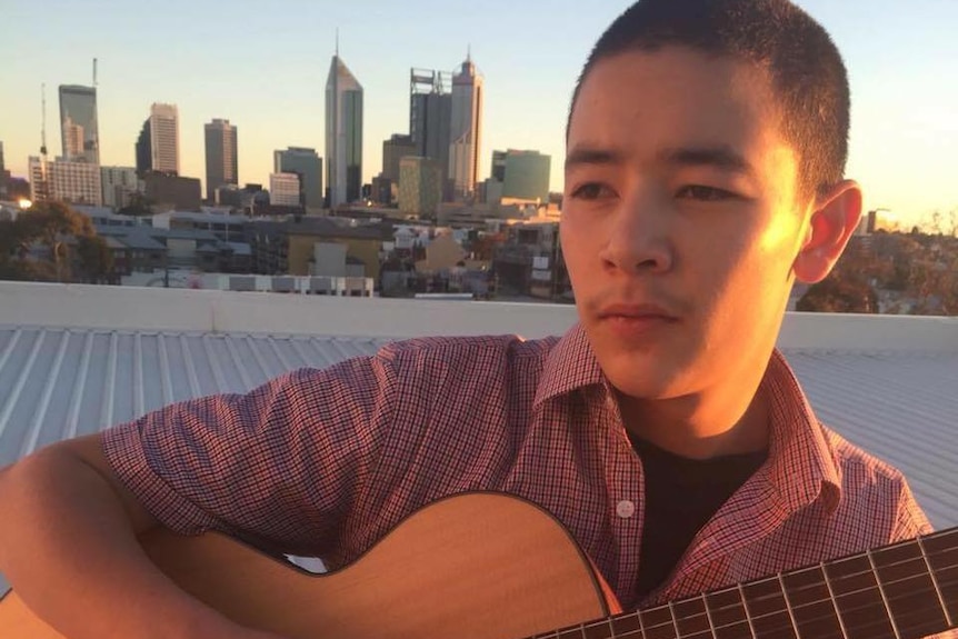 A teenager sits with an acoustic guitar with the Perth skyline behind him.