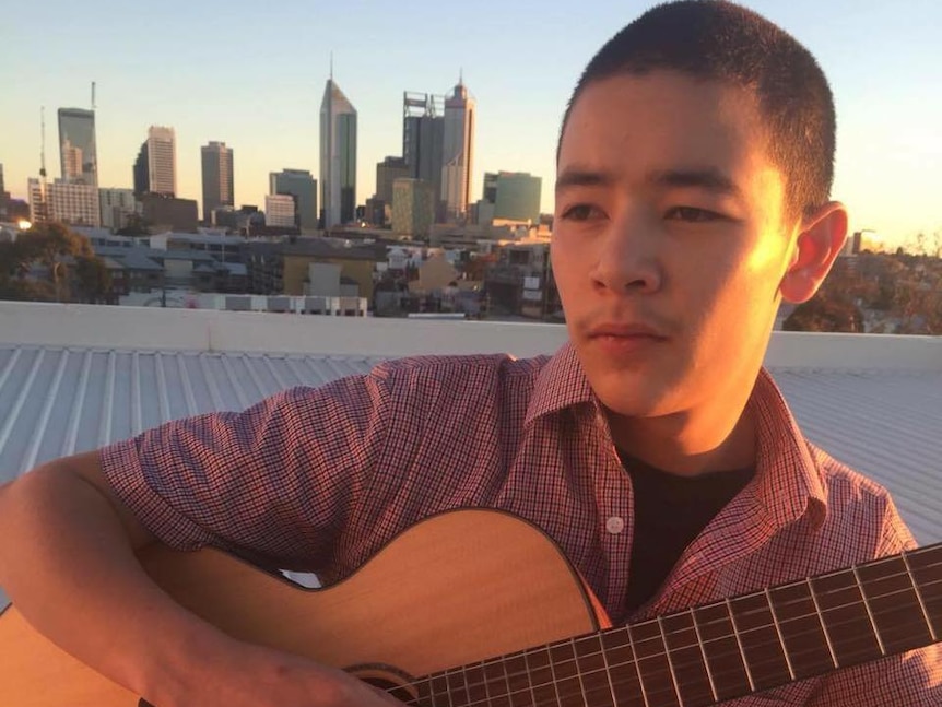 A teenager sits with an acoustic guitar with the Perth skyline behind him.