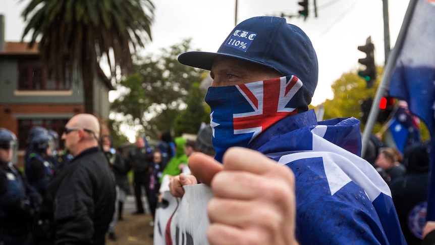 A man with the Australian flag draped across his face and around his body holds a banner and a fist to the camera at a protest