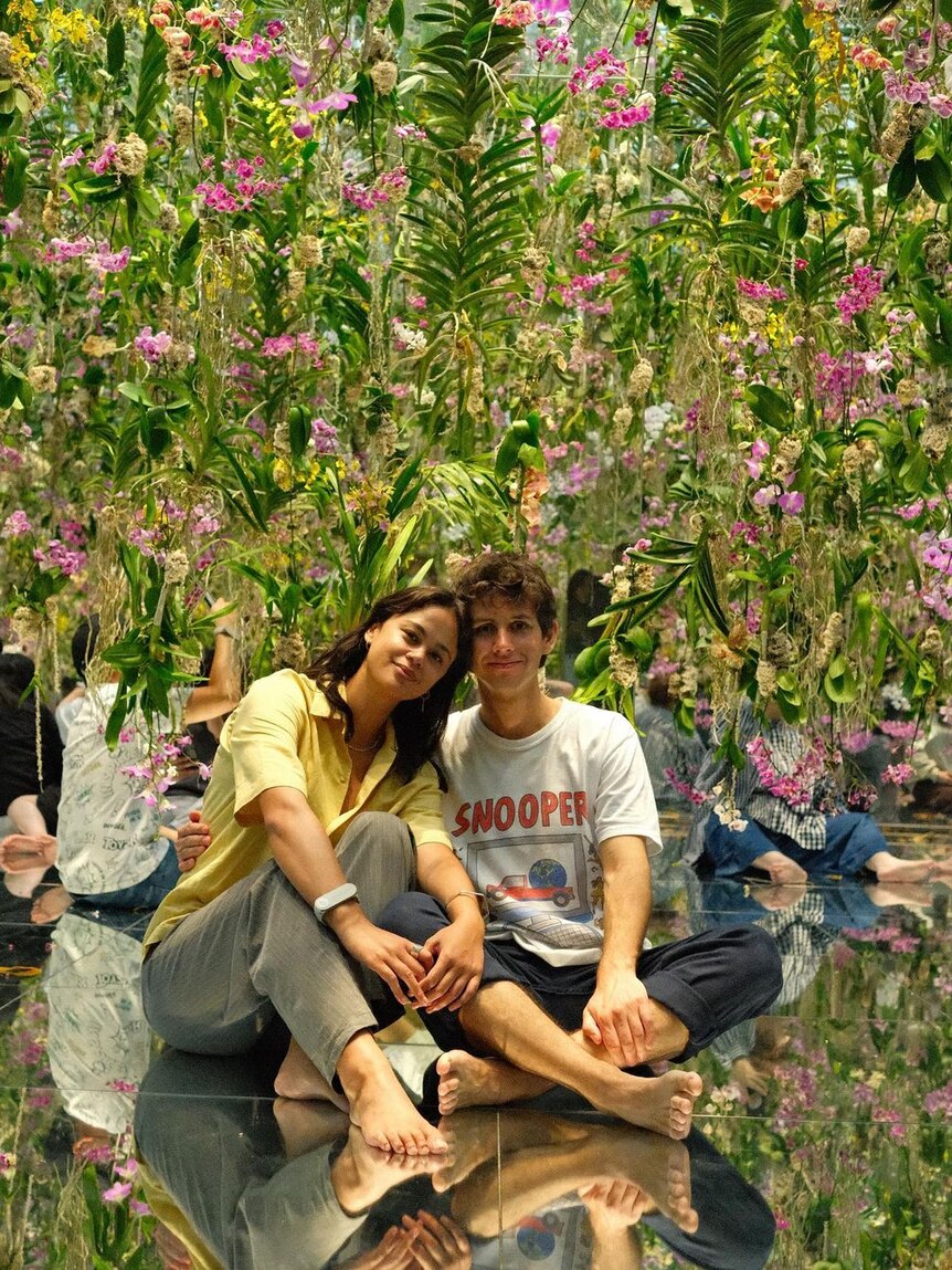 A brunette woman and man, in their 20s, sit in front of flowers and smile, heads touching. 
