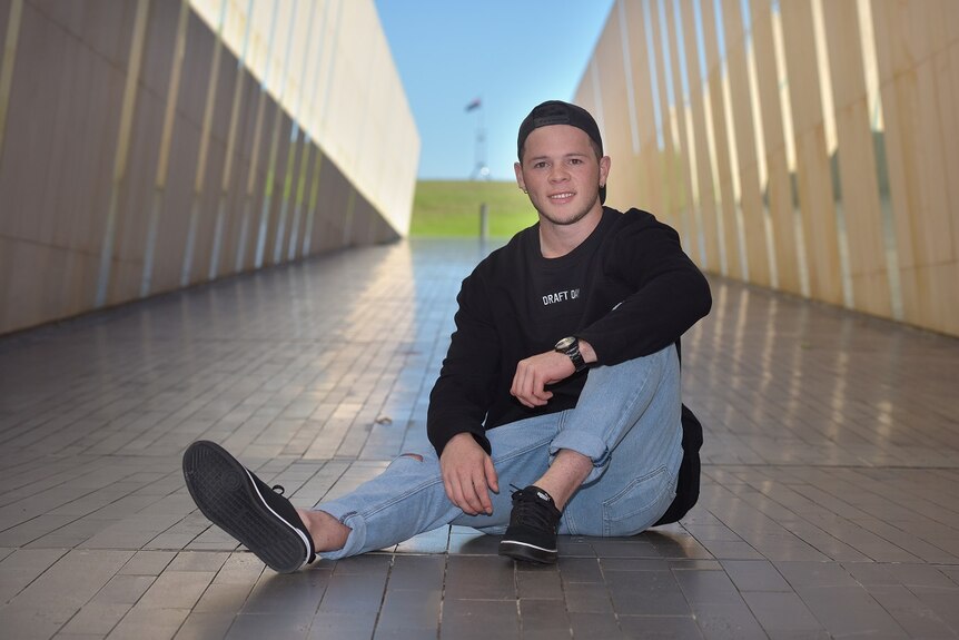 A young man in a black jumper, cap and jeans is sitting in a corridor with Parliament House in the background