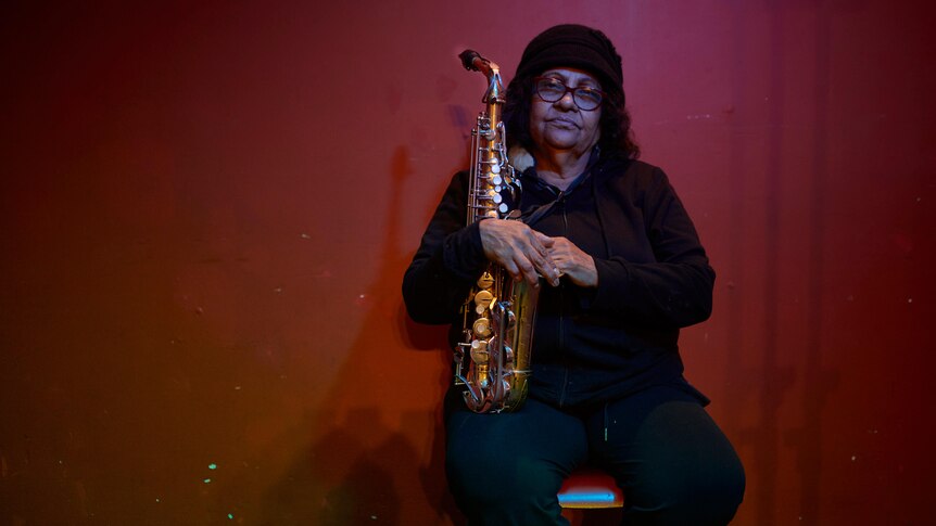 Aboriginal musican Marlene Cummins sits against red wall with saxophone looking pensive