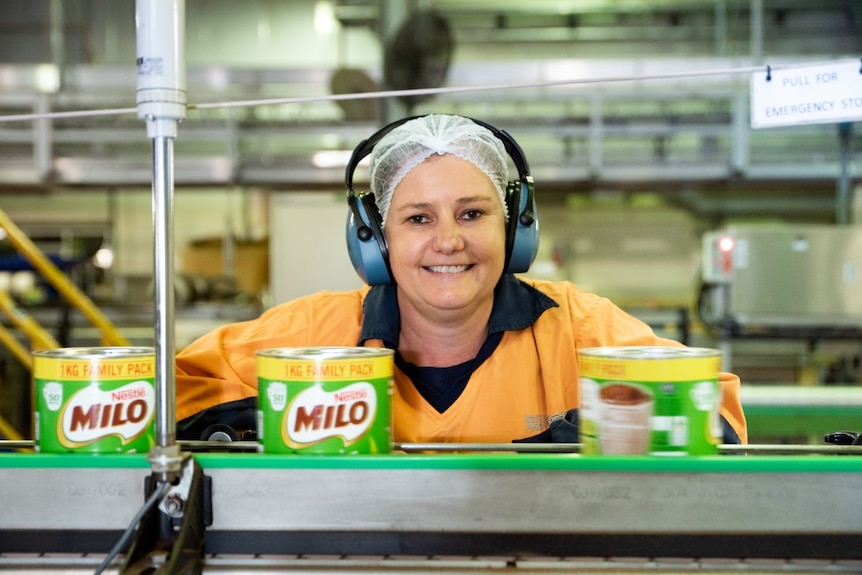 A woman wearing a hairnet and industrial earphones smiles as Milo tins are in the foreground