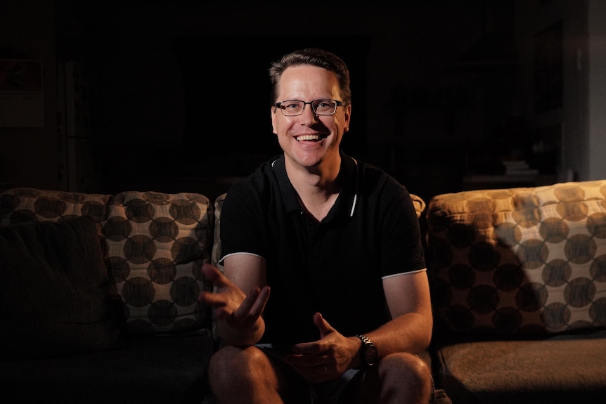 A man wearing a black polo shirt smiles at the camera. He sits on a coach and his hands are moving.