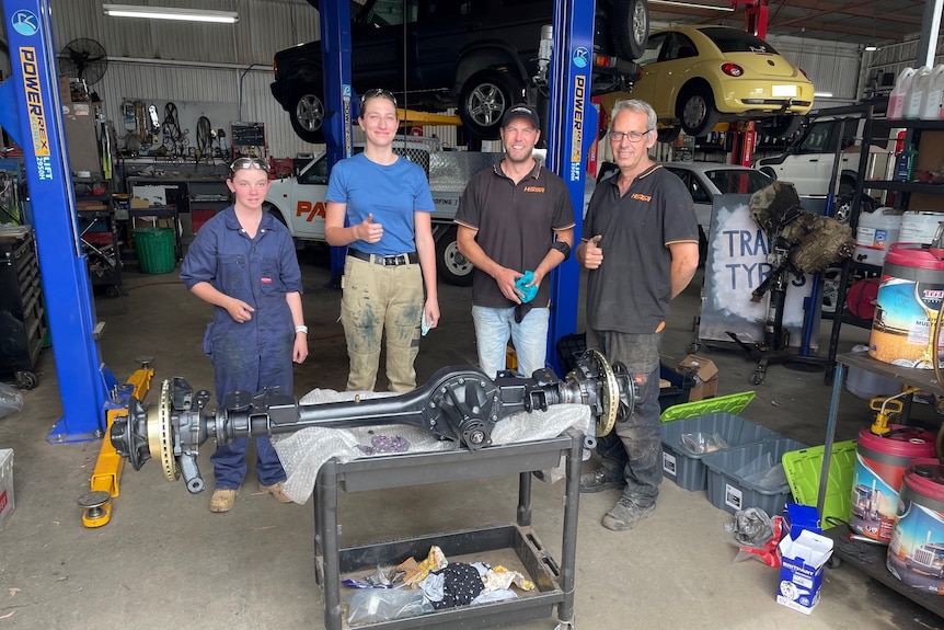 Two teen girls and two men standing in front of a car axle in a workshop.