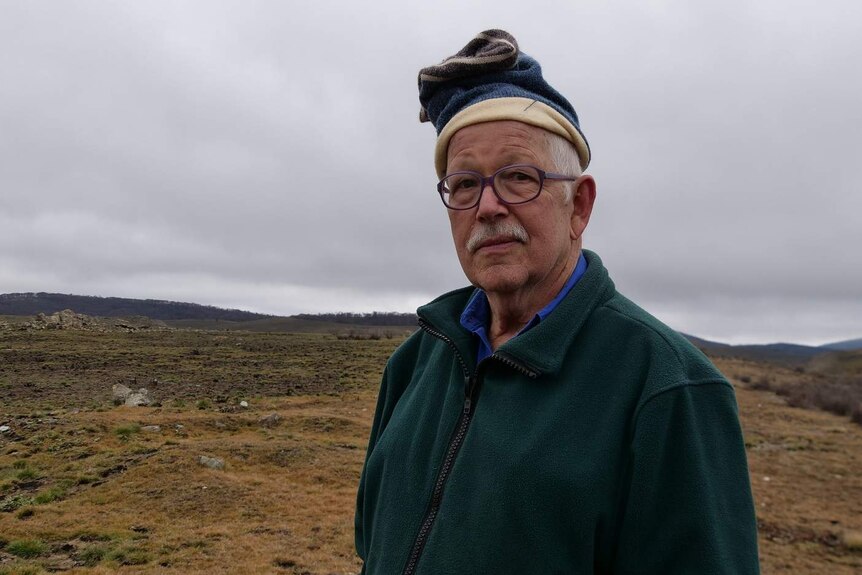An older man wearing purple glasses and a beanie, standing on a plain in Kosciuszko National Park.
