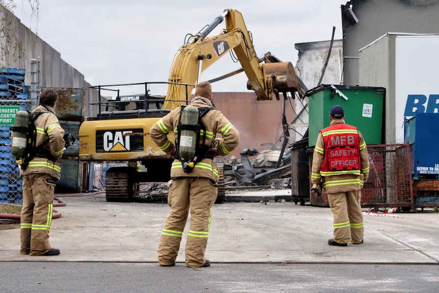 Three firefighters look on as an excavator is operated at the scene of the fire.