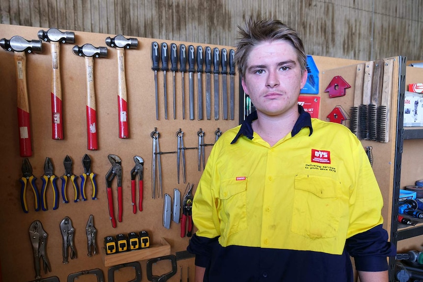 A young man stands in a workshop in front of a rack of tools.