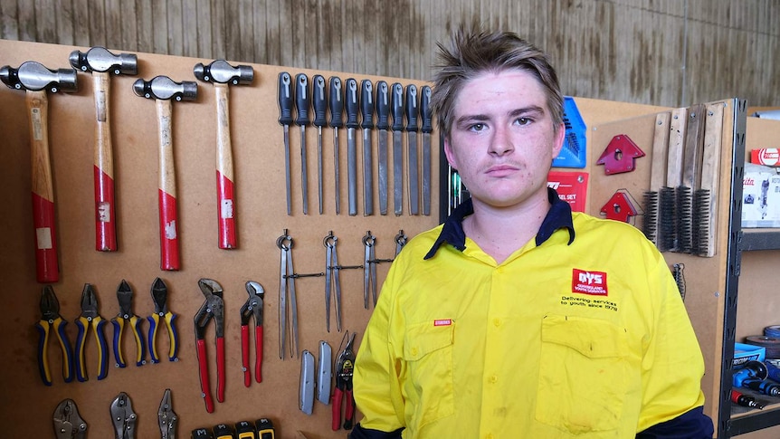 A young man stands in a workshop in front of a rack of tools.