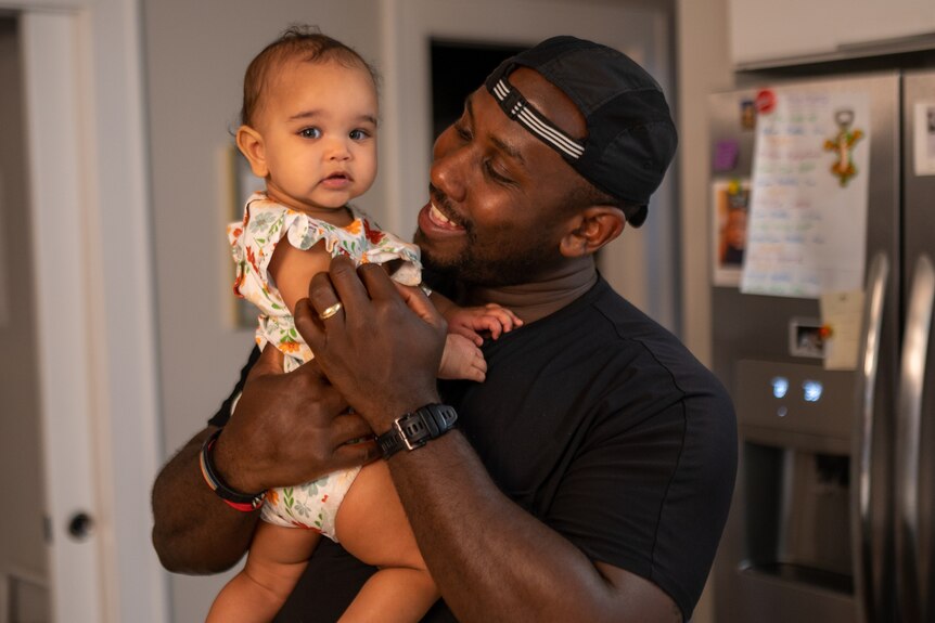 An African American man in a black T-shirt smiles while holding his infant daughter who is dressed in a floral jumpsuit