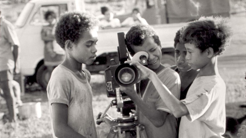 One boy looks through the viewfinder of a film video camera on a tripod and another has a hand on the lens, two others watch.
