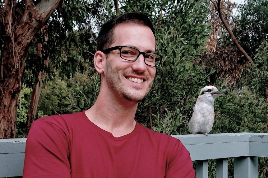 A man in glasses and a red t-shirt smiles next to a kookaburra with gum trees in the background.