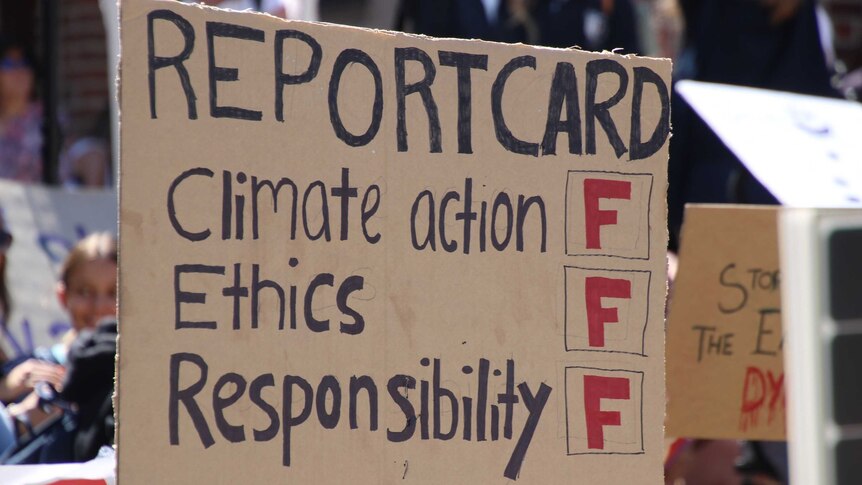 A cardboard protest card delivers a 'report card' of F on 'climate action, ethics and responsibility'.