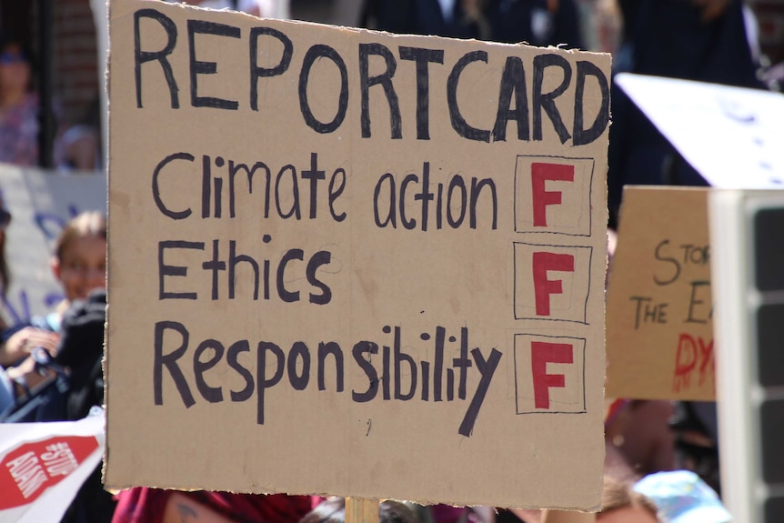 A cardboard protest card delivers a 'report card' of F on 'climate action, ethics and responsibility'.