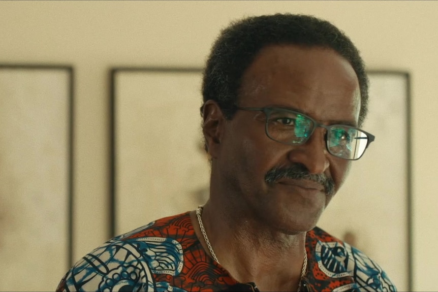 An older Black man with glasses and a moustache stands inside a room, where seemingly destroyed paintings hang on the wall
