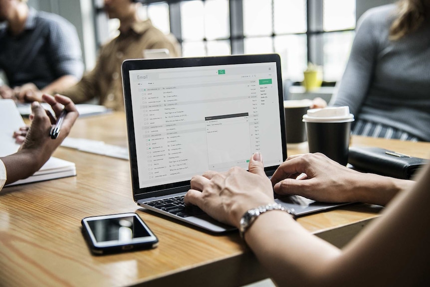 A generic image of a woman checking emails while at a table of work colleagues.