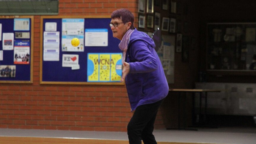 A 70-year-old woman with purple hair prepares to hit a yellow ball with a paddle while playing pickleball