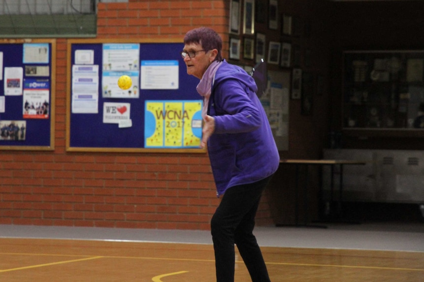 A 70-year-old woman with purple hair prepares to hit a yellow ball with a paddle while playing pickleball