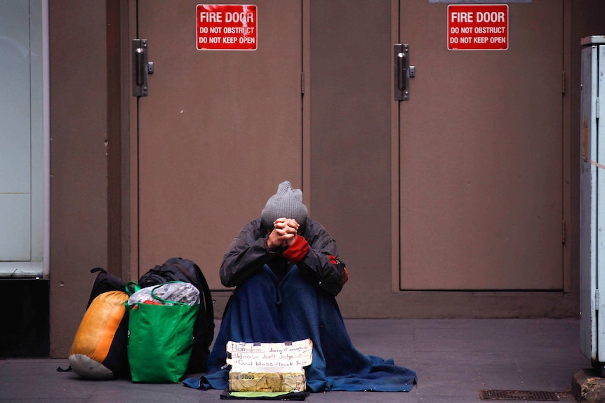 A person sits on the street with bags next to them begging for money with hands clasped together in front of their face.