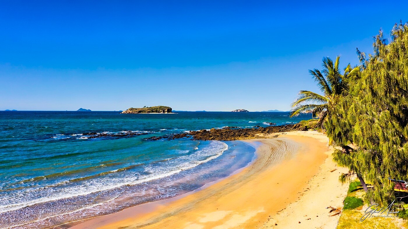 A sunny shoreline with small waves lapping on sand next to palm trees, island in distance