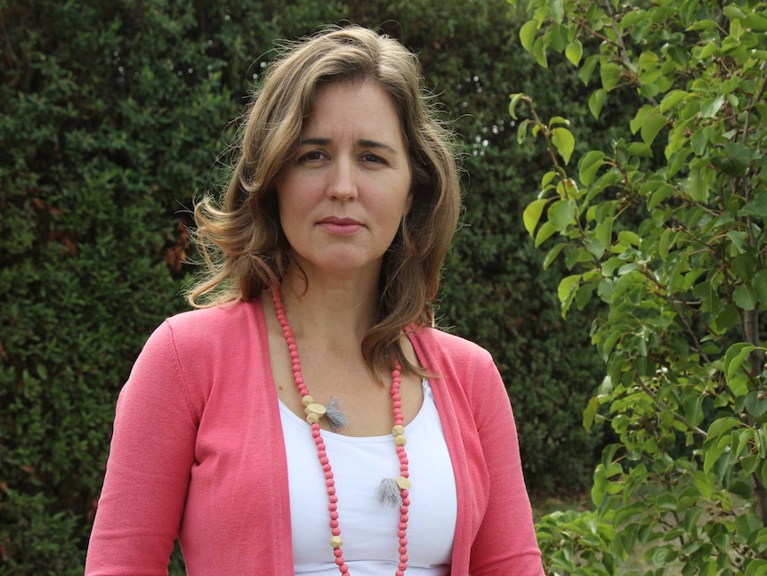 A woman in a pink cardigan in front of green trees looking at the camera with a serious expression.