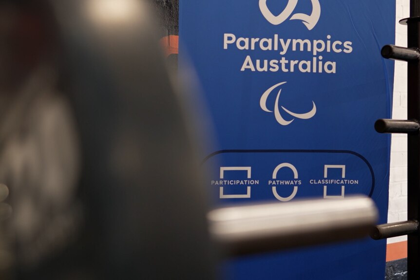 A sign that reads 'Paralympics Australia' has its logo and the Paralympic Games logo. Out of focus in the foreground is a weight