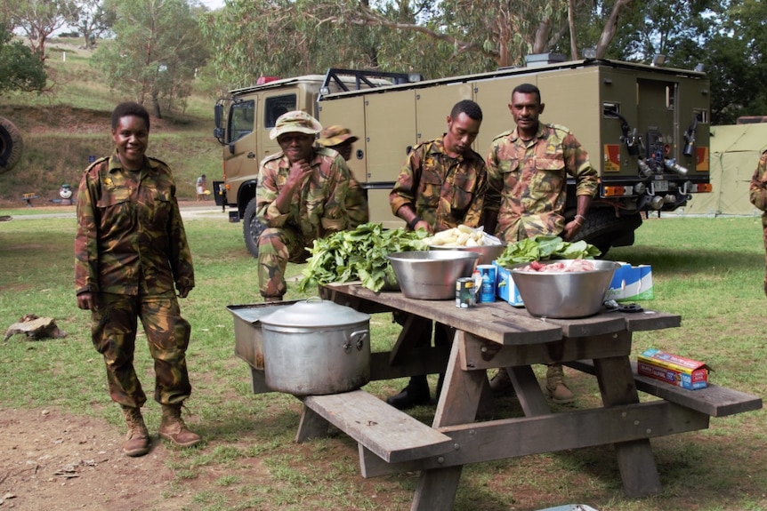 A woman in army camouflage stands next to tree men in front of a table with large bowls full of food to prepare.