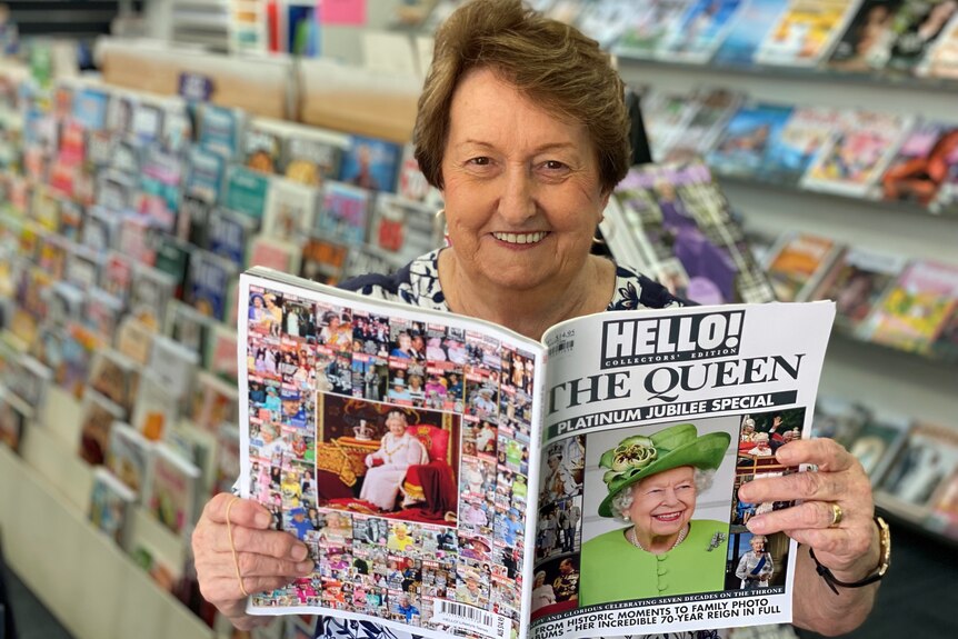 Woman sitting in newsagents holding up a magazine with Queens Elizabeth on cover.