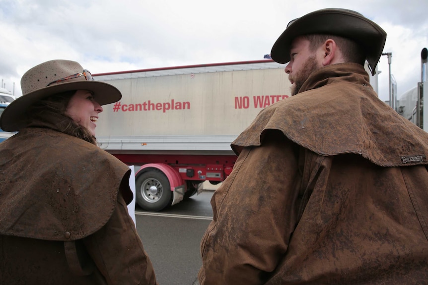 A man and a woman, both dressed in Drizabone coats, stand near a semi-trailer that says 'Can the Plan'.