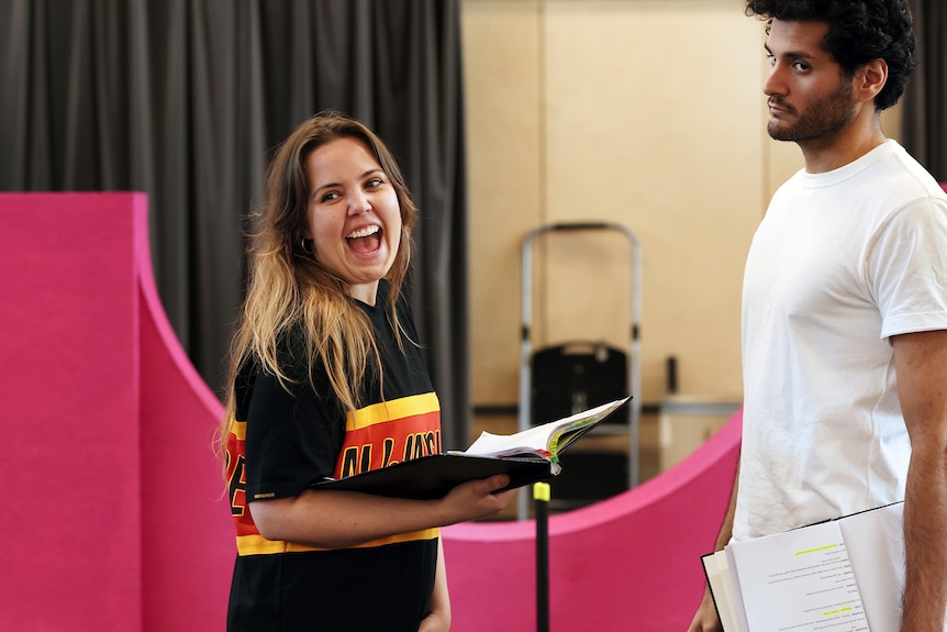A young white woman in her 30s holds a rehearsal script and laughs beside a man of colour in a white shirt.