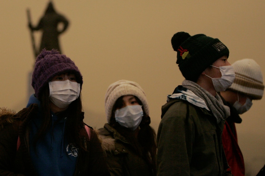 Three people wear masks against a dusty orange sky