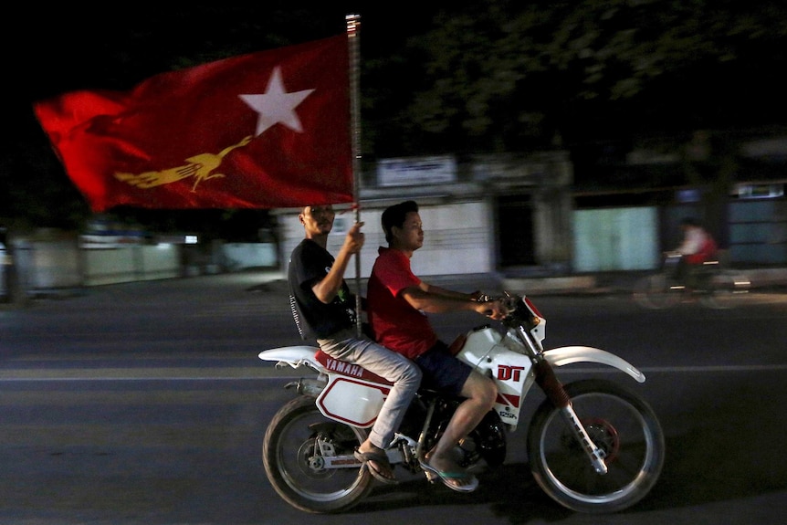 Two supporters of the National League for Democracy ride a motorbike at night while carrying a big flag.